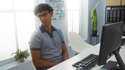 Young man wearing glasses in a modern office setting, sitting thoughtfully at a desk with a computer, potted plant, and office supplies visible in the background.