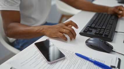 Fototapeta premium Young man working at a desk in an office using a computer keyboard with documents and a smartphone nearby