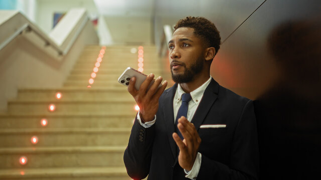 Young businessman talking on smartphone indoors, standing beside staircase, wearing suit and tie
