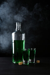 Absinthe in shot glasses, bottle and brown sugar on table against black background with smoke