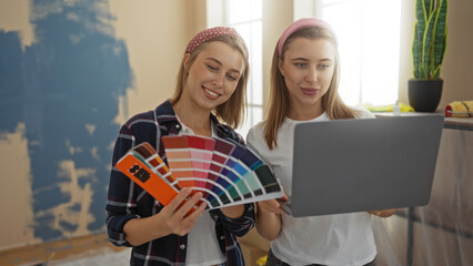 Sisters choosing colors for home renovation on a laptop while smiling and holding paint swatches in a cozy living room highlighting family and creativity.
