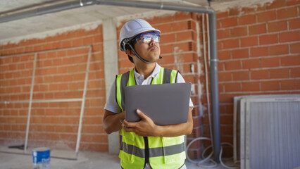 Young chinese man holding a laptop at a construction site indoors, equipped with a hard hat and safety vest, inspecting the building structure.
