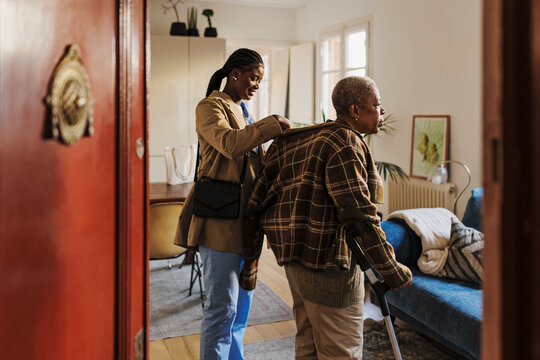 Caregiver helping elderly woman with walking stick putting on jacket