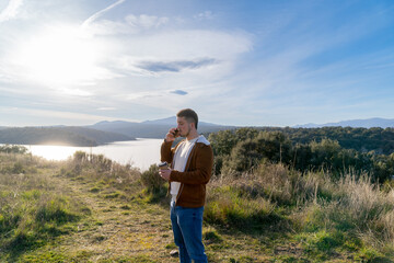 Young man talking on phone and drinking coffee in nature with lake view