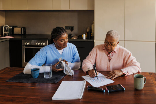 Caregiver drying dishes while senior woman is coloring mandalas