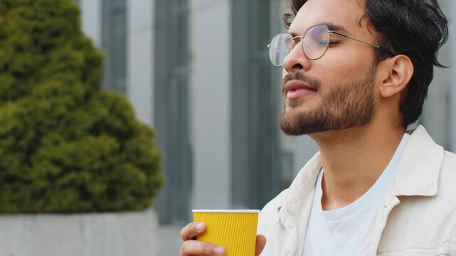 Happy young Indian man enjoying morning coffee hot drink and smiling outdoors. Relaxing, taking a break. Arabian Hindu guy in urban city downtown street, drinking coffee to go. Town lifestyles outside