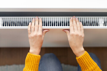Woman warming hands near modern heating radiator in room at home, above view, closeup
