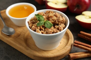 Delicious apple crisp in bowl, fresh fruits, cinnamon sticks, honey and mint on grey table, closeup