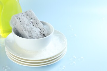 Sponge with foam, stack of dishware and bubbles on light blue background, closeup. Space for text