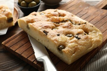 Piece of delicious focaccia bread with olives, thyme, salt and knife on wooden table, closeup