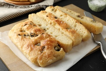 Slices of delicious focaccia bread with olives, thyme, salt and knife on black table, closeup