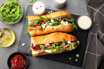 Halves of baguette sandwich with prosciutto and sun dried tomatoes and ingredients on grey table, flat lay