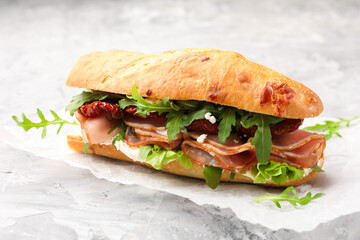 Baguette sandwich with ham and sun dried tomatoes on grey textured table, closeup
