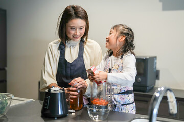 A Japanese parent and child, a woman in her 30s, a man in his 20s, and a 6-year-old girl, are making juice by putting cut carrots and honey in a blender in their home kitchen.
