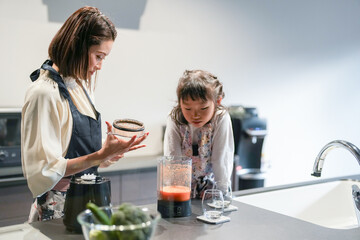 A Japanese parent and child, a woman in her 30s, a man in his 20s, and a 6-year-old girl, are making juice by putting cut carrots and honey in a blender in their home kitchen.