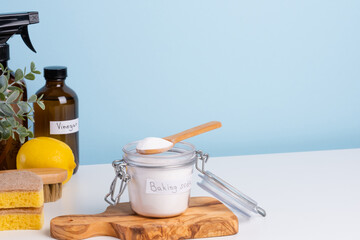 A jar filled with baking soda and a wooden spoon rests on a countertop alongside a bottle of vinegar, fresh lemon, brush and cleaning sponges
