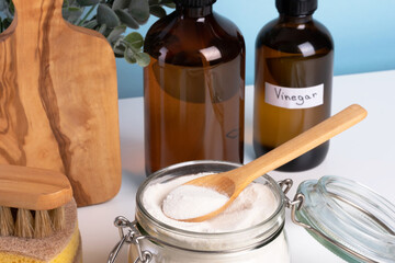 A jar filled with baking soda and a wooden spoon rests on a countertop alongside a bottle of vinegar and cleaning sponges. DIY cleaning method using natural products to remove stains. top view 