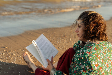 Young woman sitting barefoot on warm beach sand, immersing herself in a captivating book during a...