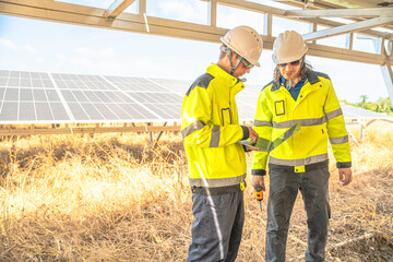 Two solar technicians in bright reflective jackets and helmets are inspecting the wiring under solar panels. One holds laptop for data logging, while the other examines electrical connections closely.