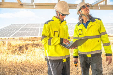Two solar technicians in bright reflective jackets and helmets are inspecting the wiring under solar panels. One holds laptop for data logging, while the other examines electrical connections closely.