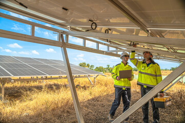 Two technicians in high visibility jackets and helmets are inspecting the underside of solar panels at a solar farm. One holds a drill, while the other uses a laptop for analysis and reporting.