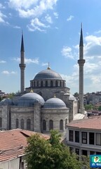 Naklejka premium In the center of Istanbul, with a blue sky and white clouds in the background, there is an ancient mosque building with domes on top. The buildings around it have dark gray roofs. On one side, you can