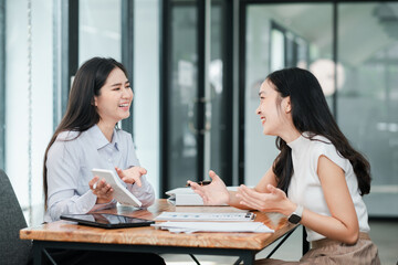 Two women in a modern office setting, discussing work with digital devices and documents on the table.