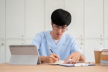 A smart Asian male medical student in scrubs, focused on studying at his desk in a classroom.