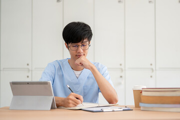 A smart Asian male medical student in scrubs, focused on studying at his desk in a classroom.