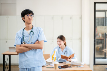 A confident Asian male medical student in scrubs is standing with his arms crossed in the classroom.