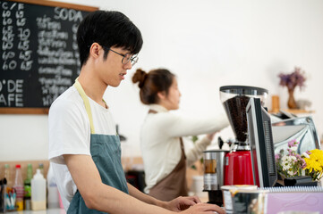 A friendly Asian male barista is working on a computer at the cashier checkpoint at the counter.