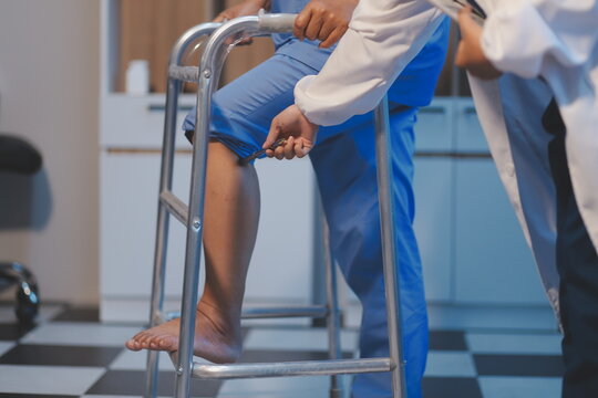 Asian young professional successful friendly female nurse in blue hospital uniform helping supporting physical therapy senior old pensioner unhealthy injury woman patient walking via four legs walker.