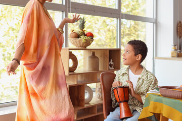 African-American boy playing tam-tam drum with his dancing mother at home. Kwanzaa celebration