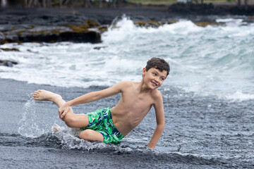 Teenager boy in colorful swimming trunks is sitting at the water's edge on the black sand, mossy rocks on the background. Black sand beach, Big Island, Hawaii, USA.