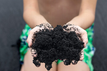 A teen boy in vivid swimming trunks is sitting in the beach and holding a handful of unique beautiful black volcanic sand made of lava. Black sand beach, Big Island, Hawaii, USA.