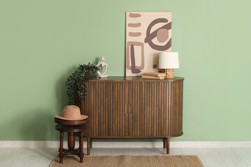 Chest of drawers with lamp, picture and hat on stool near green wall in room