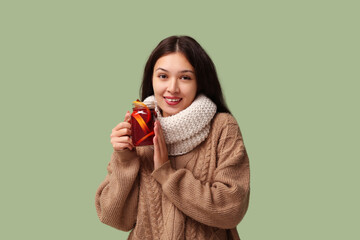 Beautiful young happy Asian woman in winter clothes with glass jar of hot mulled wine on green...