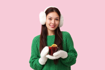 Beautiful young happy Asian woman in winter clothes with cup of hot mulled wine on pink background