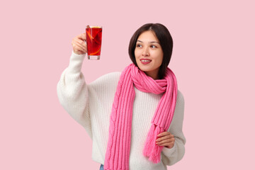 Beautiful young happy Asian woman in winter clothes with cup of hot mulled wine on pink background