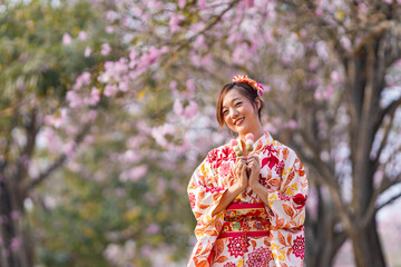 Japanese woman in traditional kimono dress holding sweet hanami dango dessert while walking inside park at cherry blossom tree during spring sakura festival