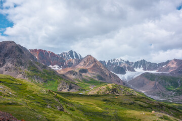 Vivid dramatic alpine scenery with green hills and rocks with view to high rocky pointy peak, sharp rockies of red color, large snow mountain top, snowy range and big glacier tongue under cloudy sky.