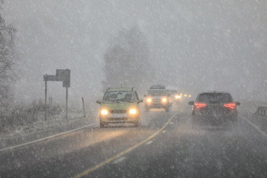 Driving in highway traffic with winter sleet and snow creating low visibility conditions, facing bright headlights of the oncoming traffic.