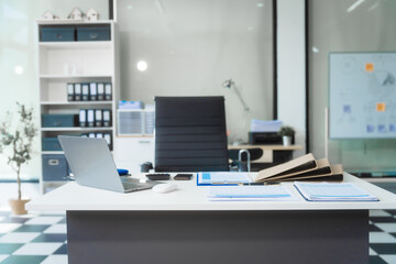A modern office room with daylight streaming in, featuring a sleek business desk, black leather chair, laptop, financial charts on A4 papers, a whiteboard, and a printer.