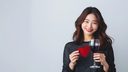 A joyful Asian woman shows a vibrant smile while holding a small red heart in one hand and a glass of red wine in the other. Single Awareness day or Valentine.