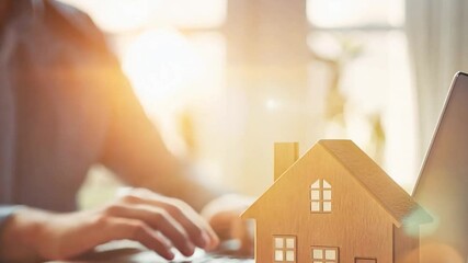 A close-up of a wooden house model with stacked coins symbolizing property investment and financial planning. A laptop and business documents in the background emphasize the digital and analytical