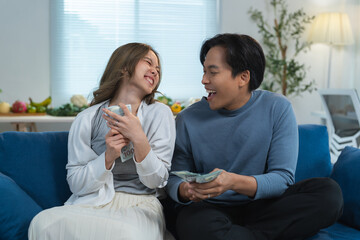 Young asian couple holding money and laughing together, celebrating financial success, lottery win, or profitable investment while sitting on a comfortable sofa in their modern living room
