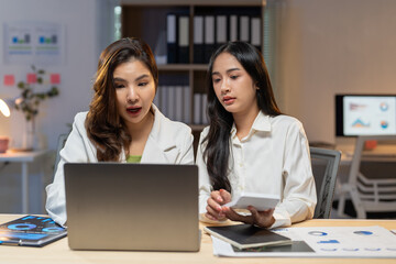 Two young asian businesswomen collaborating on a project, analyzing financial data on a laptop while using a calculator in a brightly lit office at night