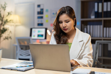 Young asian businesswoman sitting at her desk concentrating on working on her laptop, focused on her tasks in a dimly lit office late at night