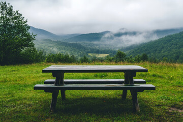 Fototapeta premium Empty wooden picnic table and bench on green meadow with beautiful view of mountains in background on cloudy summer day. Place for relax in nature