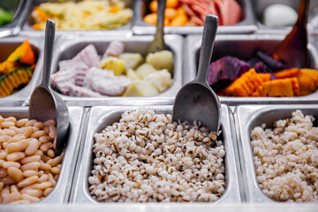 Close-up of a buffet millet, white beans, and colorful boiled vegetables, with stainless steel trays and scoops, highlighting fresh and nutritious meal ingredients.